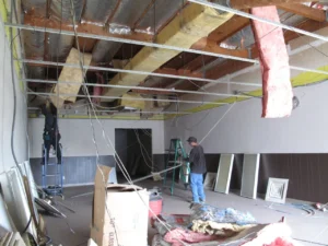 Workers remove ceiling insulation in a room with exposed framing and hanging wires.