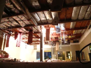 Exposed ceiling joists and insulation above a room during demolition, with dangling wires and debris below.