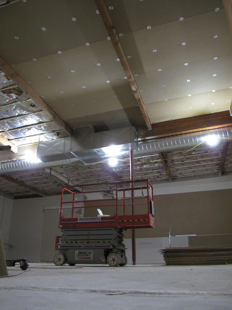 Ceiling panels cover part of the rafters above exposed ductwork, with a scissor lift and stacked materials below.