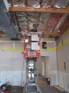 A worker on a lift removes ceiling materials beneath exposed insulation and beams in a church interior.