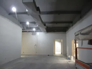 An unfinished interior room with exposed ceiling beams, bare walls, a doorway, and building materials along one side.