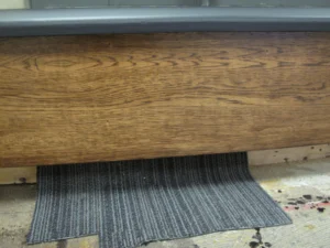A wide wooden board sits beneath a gray sill, above a striped mat and unfinished flooring.