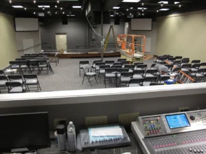 Rows of folding chairs face a raised stage as a scissor lift and audio console sit in the auditorium under construction.
