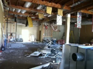 Partially demolished interior with exposed ceiling framing and insulation, metal studs, and debris across the floor.