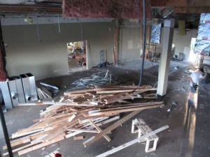 A gutted interior with exposed wiring and insulation, scattered lumber and debris, and a worker near the doorway.