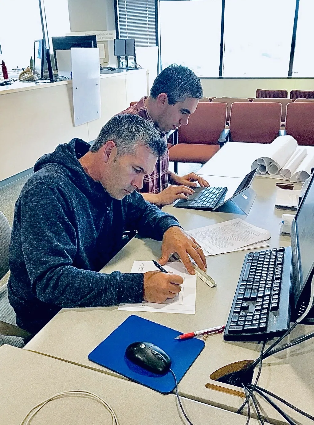 Two men review building plans at a desk; one sketches with a ruler while the other works on a laptop.